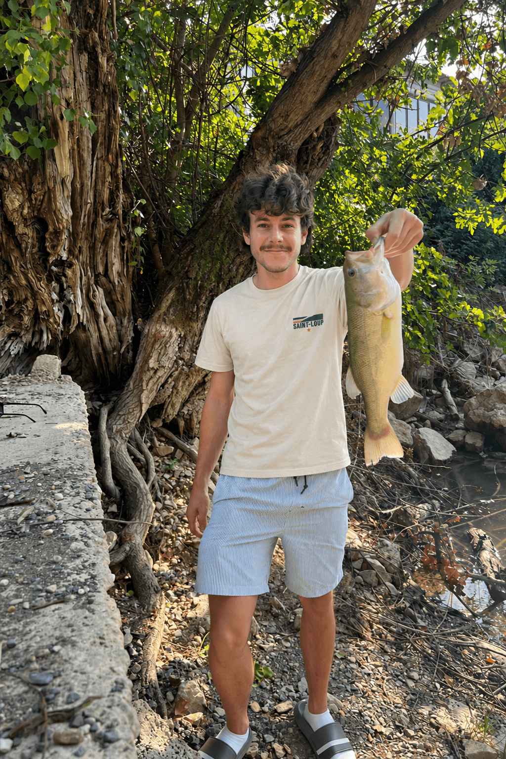 Angler wearing the Saint-Loup tee, small chest logo visible, holding a bass by the water