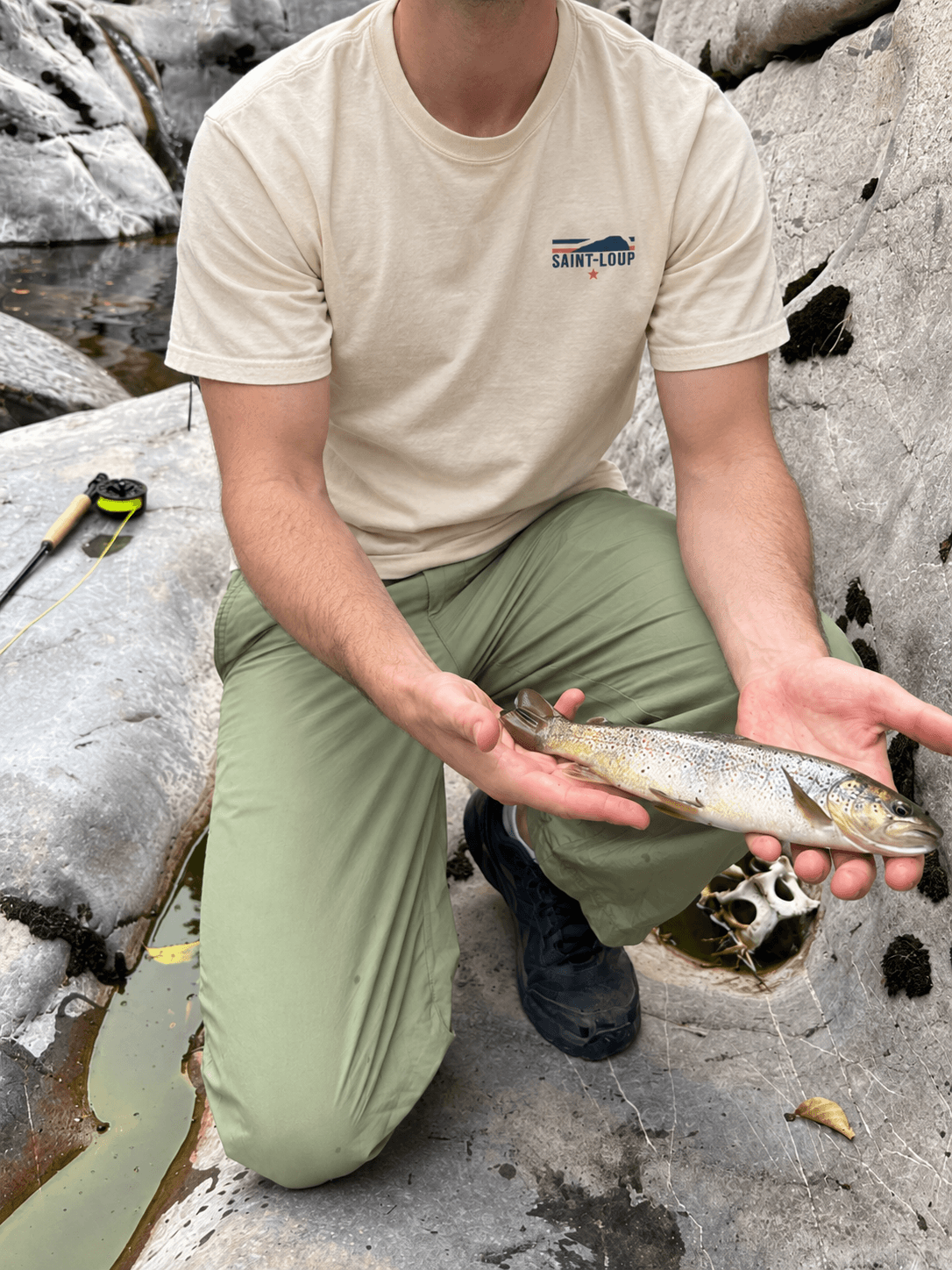 Angler wearing a Saint-Loup tee, holding a trout by the river